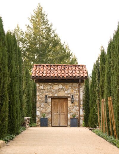 Rustic stone shed with a tiled roof flanked by tall conifer trees along a gravel pathway.