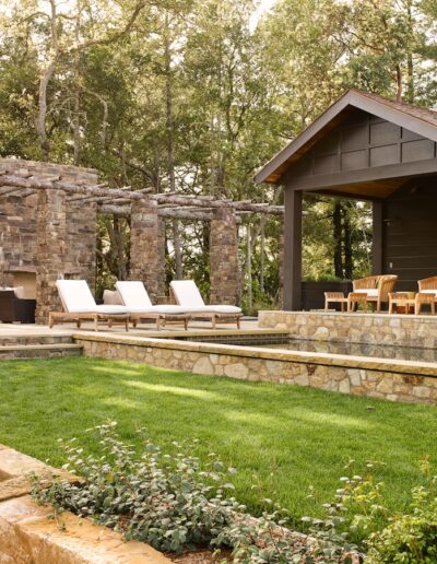Elegant outdoor living area with a pergola, stone fireplace, and comfortable lounge chairs, surrounded by lush greenery.