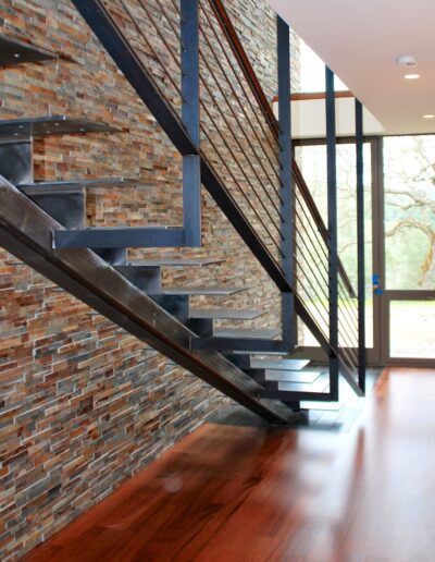Modern staircase with metal railings alongside a brick wall, leading upwards with a view of trees through large windows.