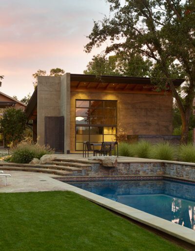 Modern backyard with swimming pool, lounging chairs, and a pool house at dusk.