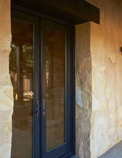A rustic patio with stone walls, a black framed glass door, wall-mounted lanterns, and a wooden rocking chair.