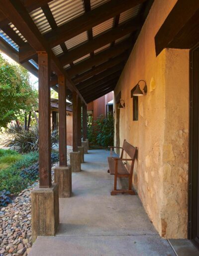 A covered walkway beside a stone wall with wooden beams and a single chair under the eaves.