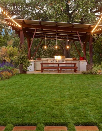 Outdoor evening setting with a pergola adorned with string lights, featuring a built-in grill and a dining area surrounded by lush landscaping.