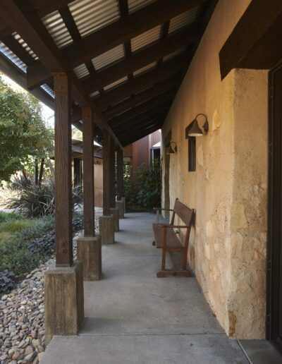 A shaded outdoor corridor featuring wooden posts, a stone wall with sconces, and a solitary wooden chair beside a landscaped garden.