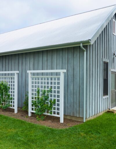 A weathered gray barn with white trellises supporting climbing plants, located in a well-manicured garden setting.