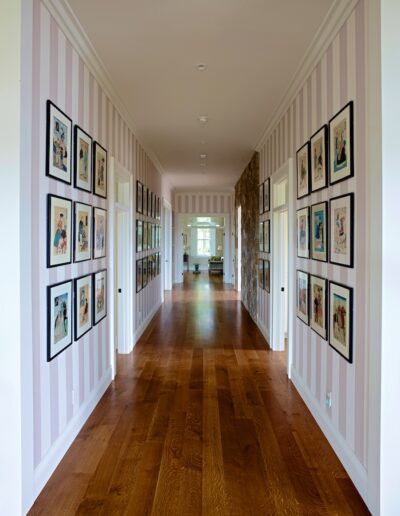 A hallway with pink-striped walls adorned with framed artworks, leading to a room in the distance.