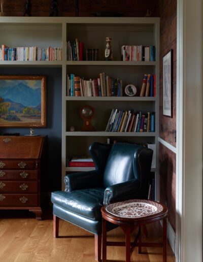 A cozy reading nook with a leather armchair, a side table, and a bookshelf, with ambient lighting and a classic painting above.