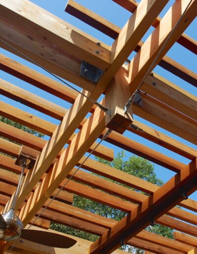 Wooden pergola structure with an outdoor fan, viewed against a blue sky.